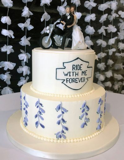 2-tier wedding cake with buttercream purple vines, a "Ride With Me Forever" plaque, and a topper with bride and groom on a motorcycle