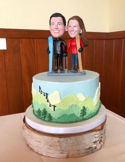 buttercream mountain scene wedding cutting cake with bobblehead bride and groom on top