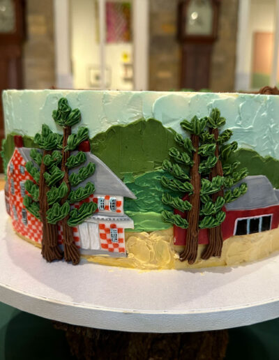 Cake with scene from Grandma Moses painting of checkered barn, buttercream trees and mountains, other barn in background.