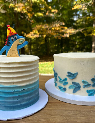 blue and white smash cake with fondant dinosaur wearing a party hat and larger white cake with blue buttercream petals
